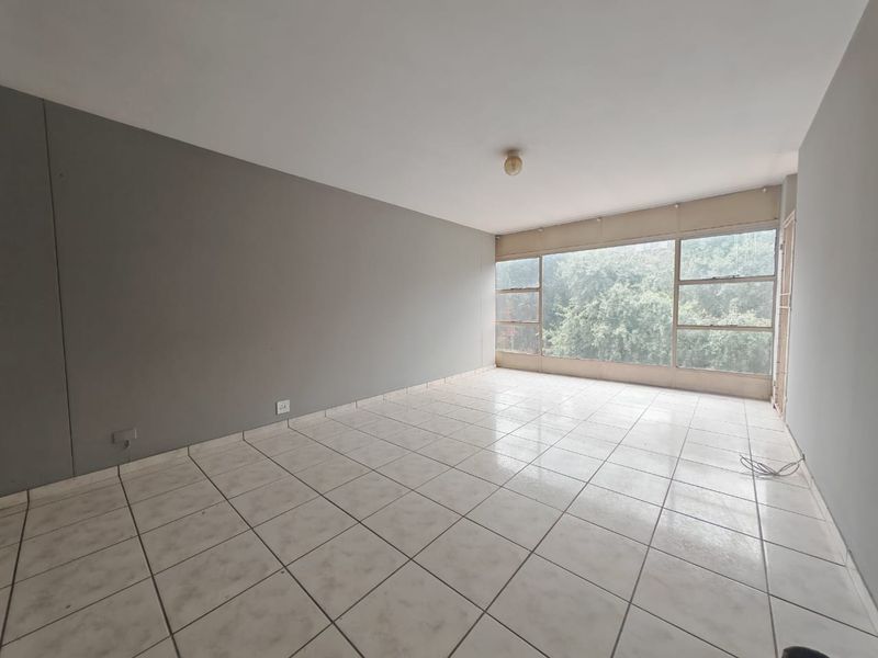 An empty, sunlit living room featuring neutral grey walls, large windows with a garden view, and tiled flooring throughout.