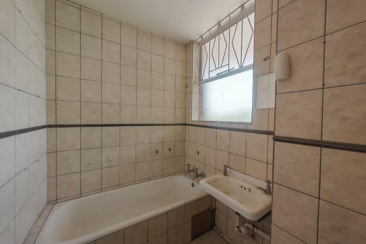 A dated bathroom featuring a standard bathtub and wall-mounted basin with full-height wall tiling and security bars on the window.