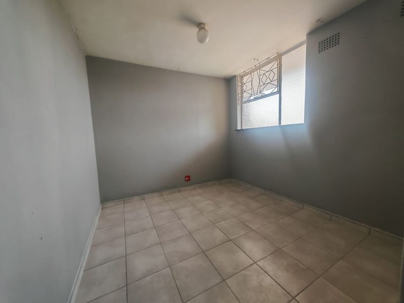 An empty bedroom featuring neutral tiled flooring, grey painted walls, and a security-fitted window, offering a blank canvas for personalization.