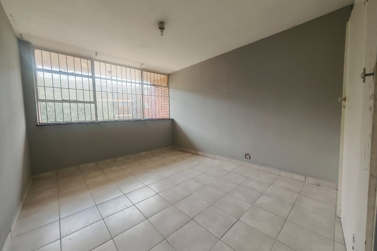 An empty bedroom featuring tiled flooring, large windows with security bars, and neutral grey walls, offering a blank canvas for renovation.