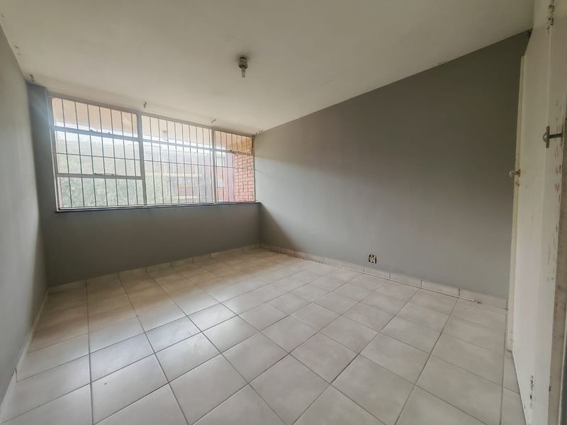 An empty bedroom featuring tiled flooring, large windows with security bars, and neutral grey walls, offering a blank canvas for renovation.