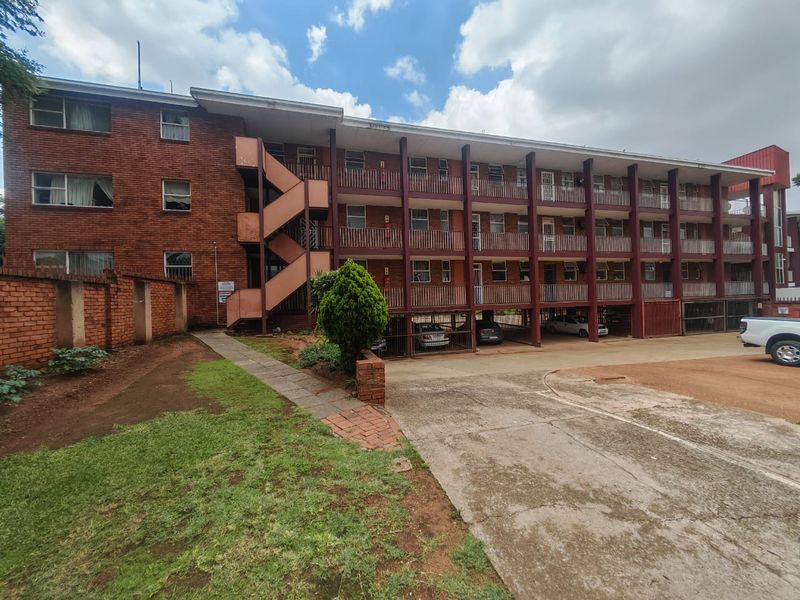 Exterior view of a multi-story brick apartment building featuring external staircases, covered parking bays, and a paved driveway area.