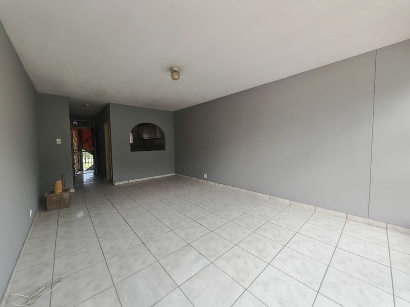 An empty, open-plan living area featuring neutral grey walls and light-colored ceramic floor tiles, with an arched opening leading to an adjacent space.