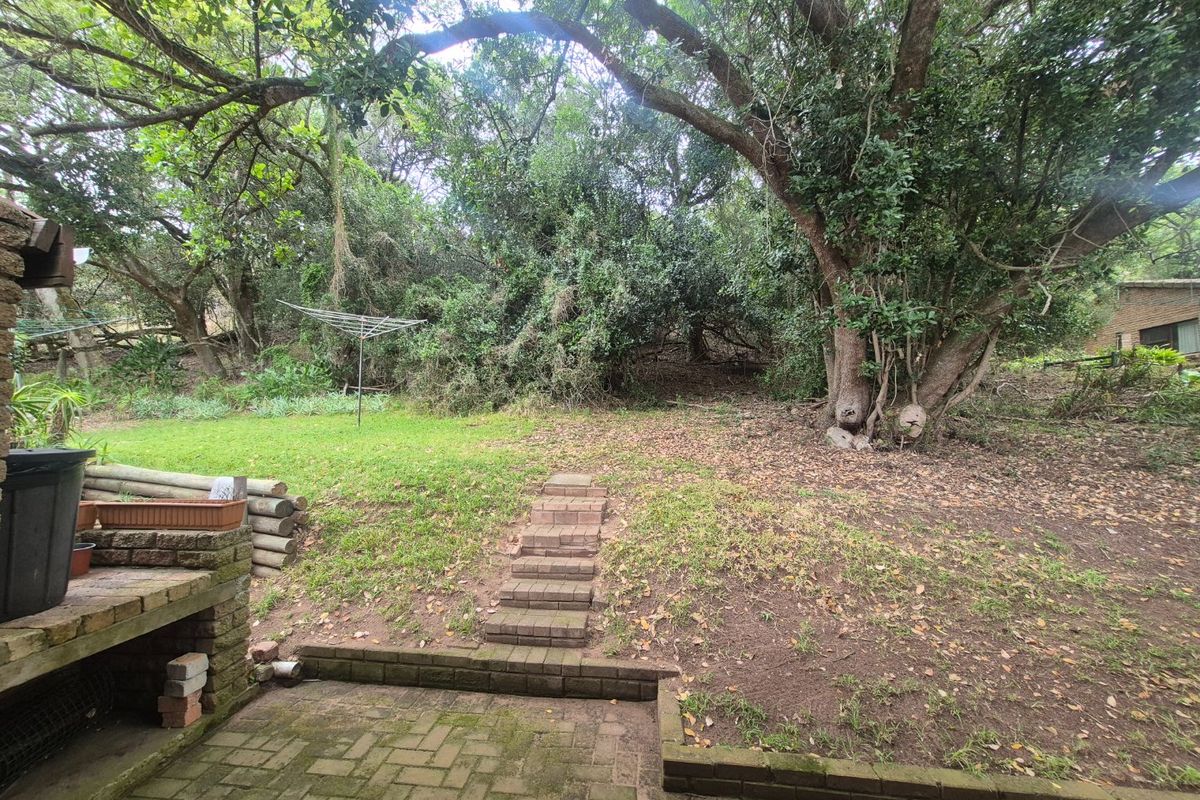 A rear garden area featuring a brick-paved patio, built-in brick braai, and tiered brick steps leading to a sloped, tree-lined lawn.
