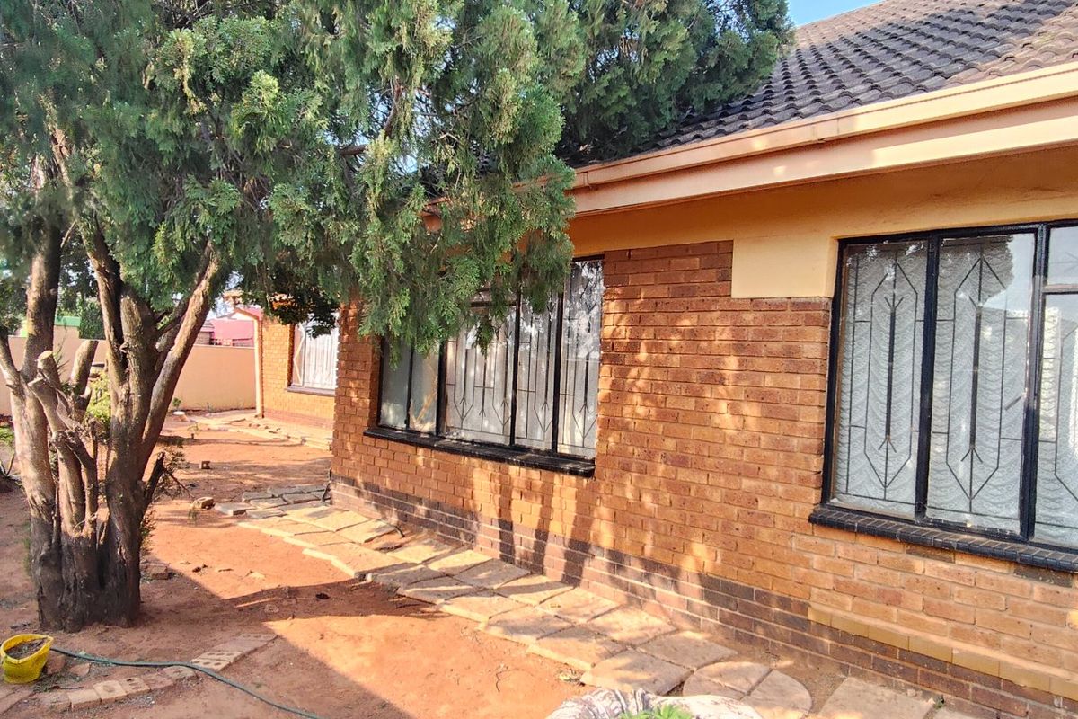 Rear view of a face brick house featuring a tiled roof, security bars on windows, and a paved garden area with mature trees.