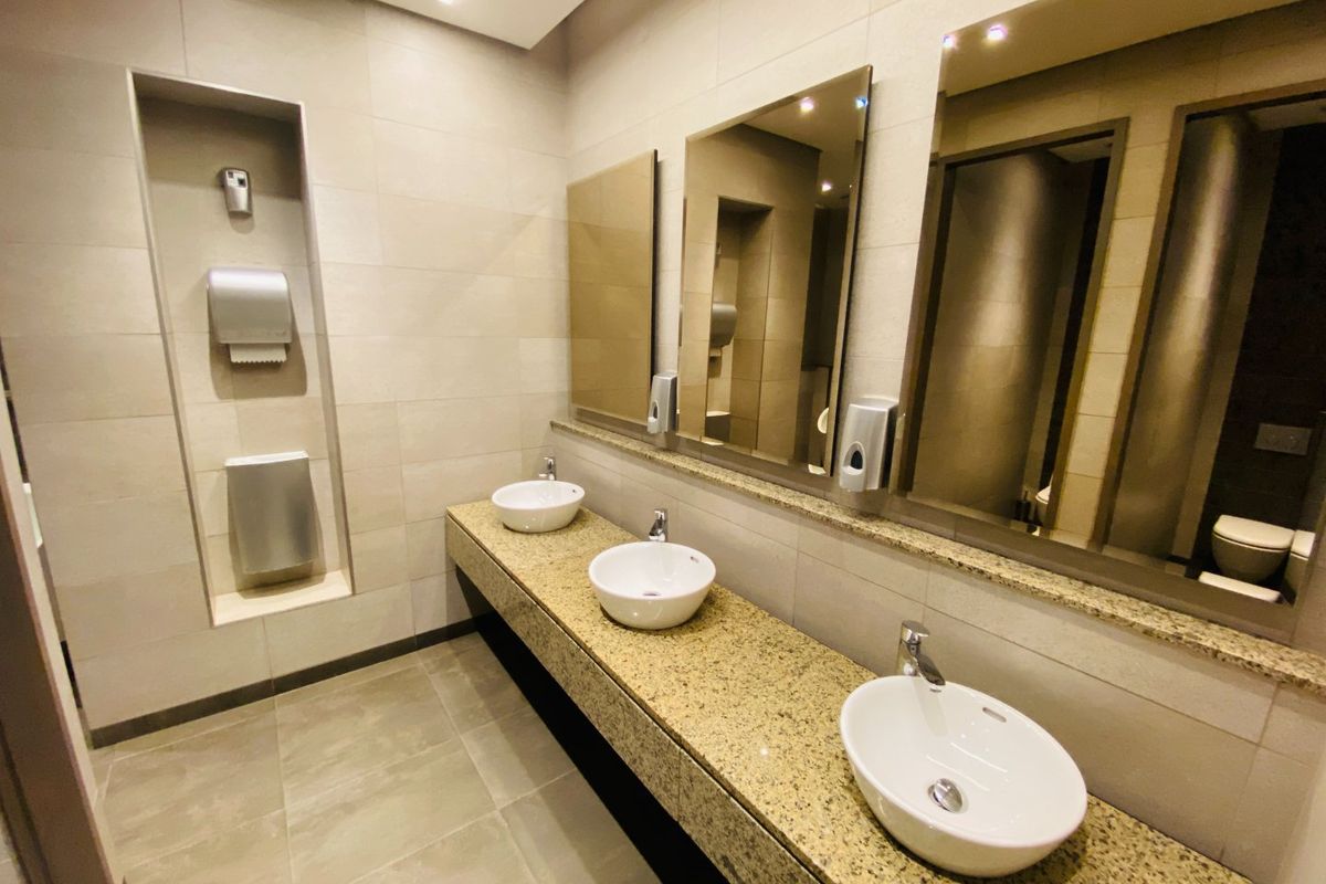 A modern commercial bathroom featuring a long granite vanity with three vessel sinks, large mirrors, and integrated wall-mounted dispensers.