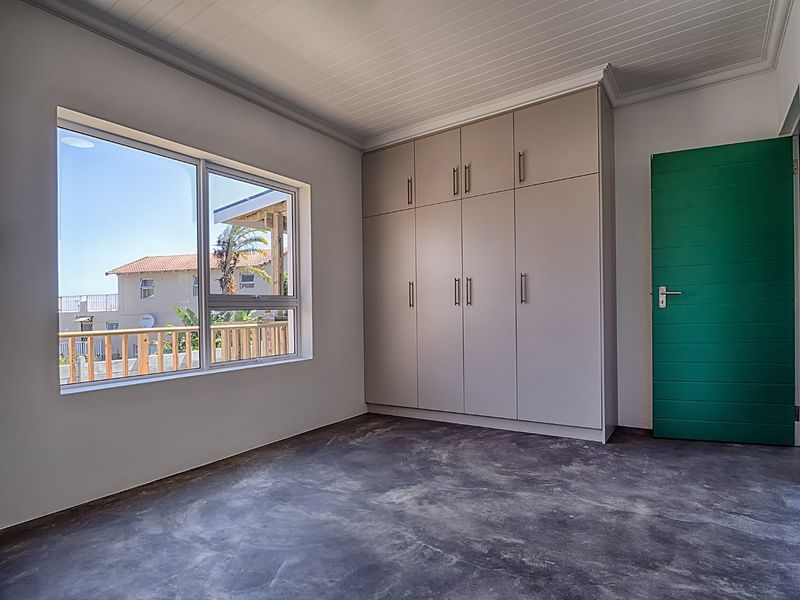 A bright, empty bedroom featuring built-in cupboards, a tongue and groove ceiling, and an unfinished concrete floor with a view of the exterior.