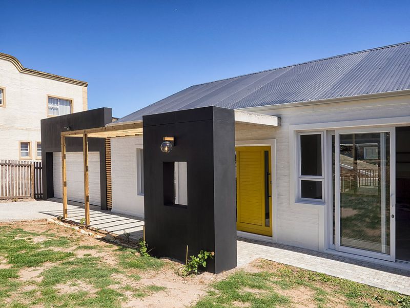 Modern exterior facade featuring a striking yellow front door, contrasting black architectural elements, a timber pergola, and a clean corrugated metal roof.