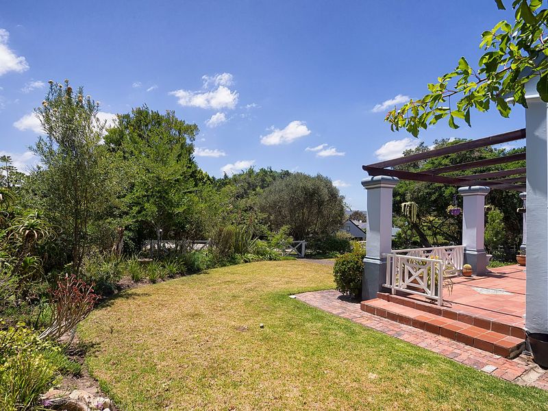 A covered patio area featuring brick paving and a pergola, overlooking a well-maintained garden with mature trees and shrubs.