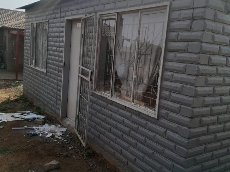 Exterior view of a single-story dwelling featuring a corrugated metal roof and security gate, situated on an unfinished plot.