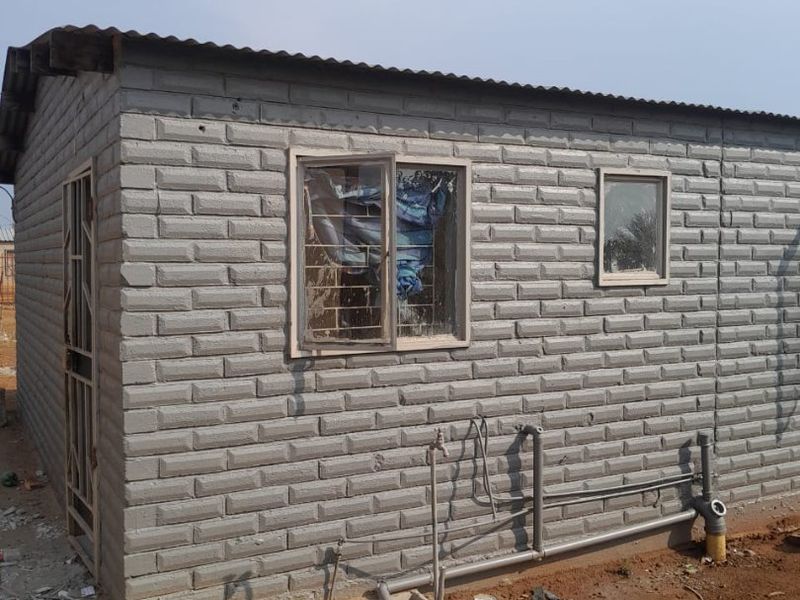 Exterior view of a basic structure featuring grey brick-style cladding, corrugated metal roofing, and exposed exterior plumbing connections.