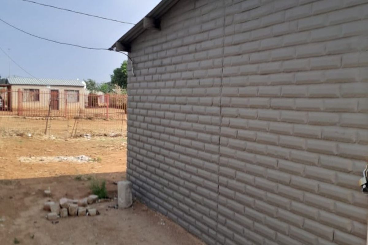 Side view of a residential structure featuring textured brick-effect cladding and a corrugated metal roof, set on an undeveloped plot.