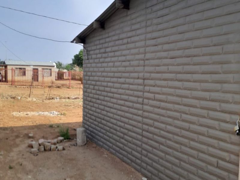 Side view of a residential structure featuring textured brick-effect cladding and a corrugated metal roof, set on an undeveloped plot.
