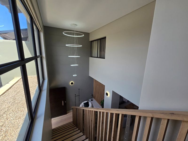 A modern, double-volume entrance hallway featuring a timber staircase, contemporary pendant lighting, and elegant timber wall paneling with large windows.