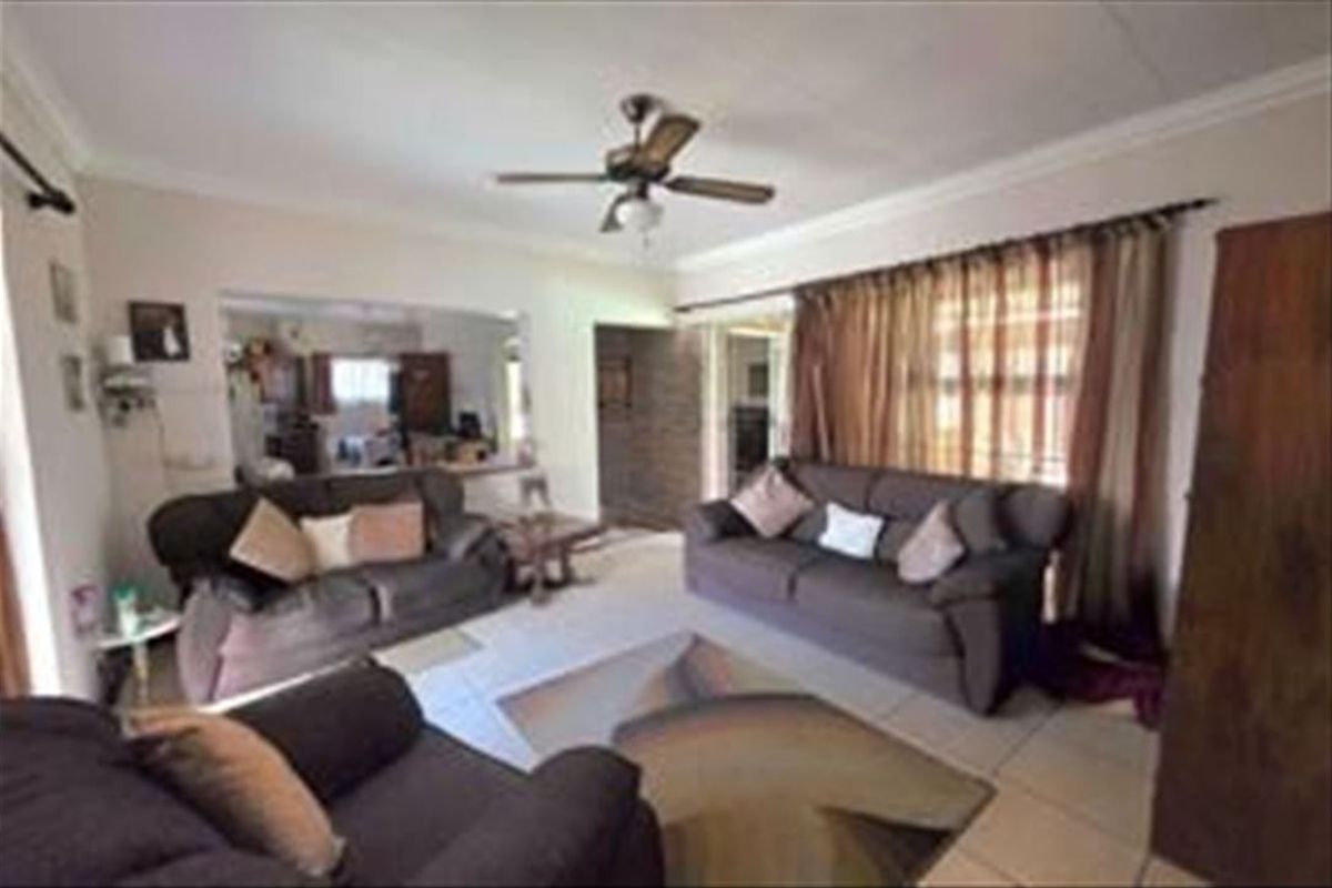 A spacious living room featuring tiled flooring, a ceiling fan, and coved cornicing, with an open-plan flow leading into the adjacent kitchen area.
