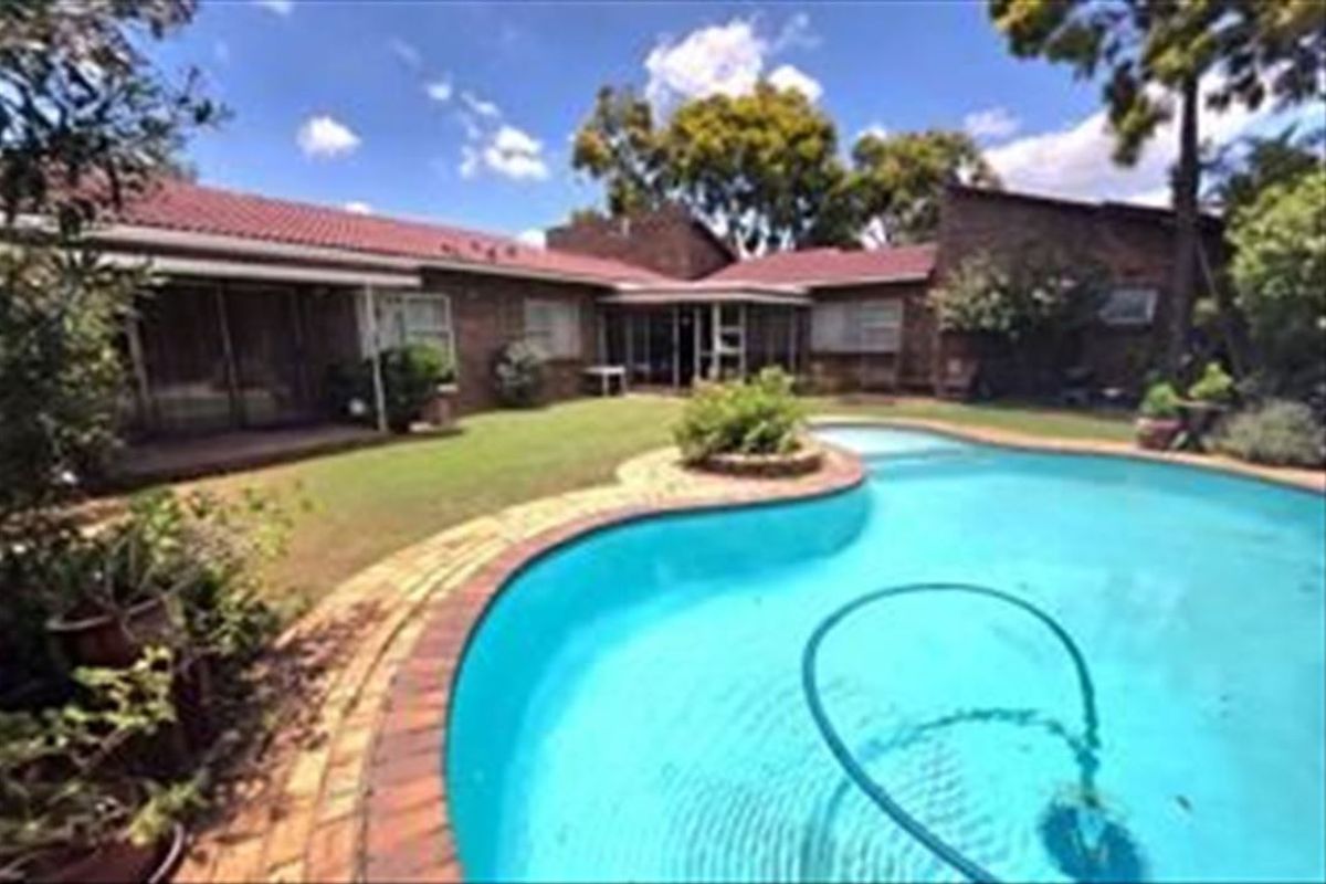 A rear view of the property featuring a swimming pool with brick paving, a landscaped garden, and a brick-built house with a tiled roof.