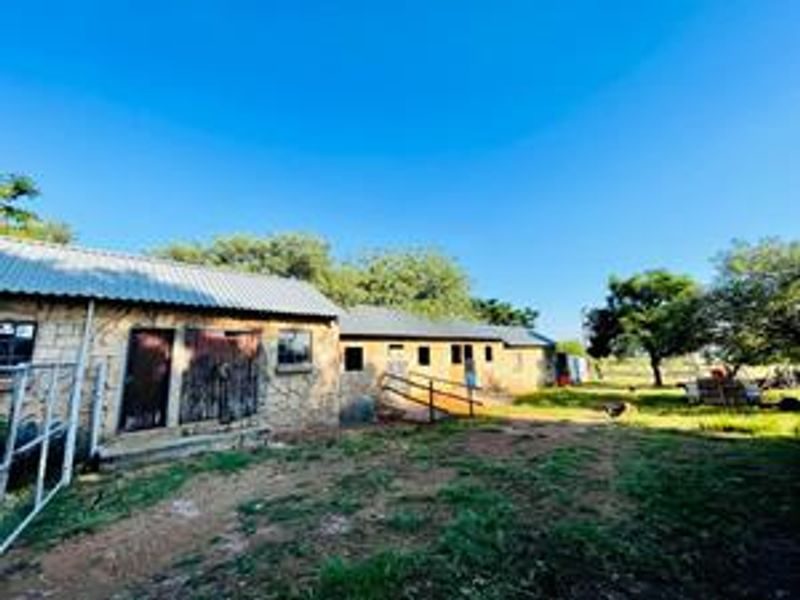 Rear view of a rustic farm building featuring stone masonry walls, corrugated metal roofing, and multiple access points set within a rural landscape.