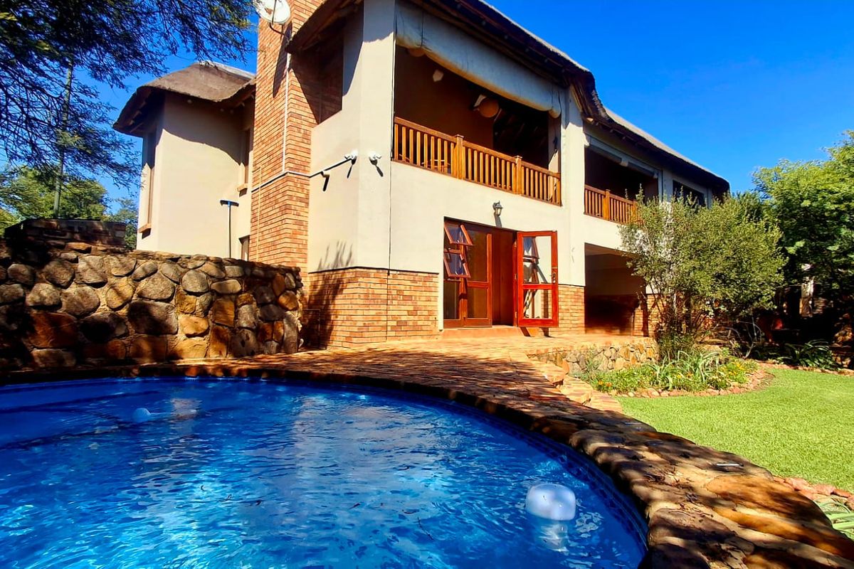 Rear view of a two-story house featuring a swimming pool, stone retaining wall, thatched roof, and a balcony with wooden railings.