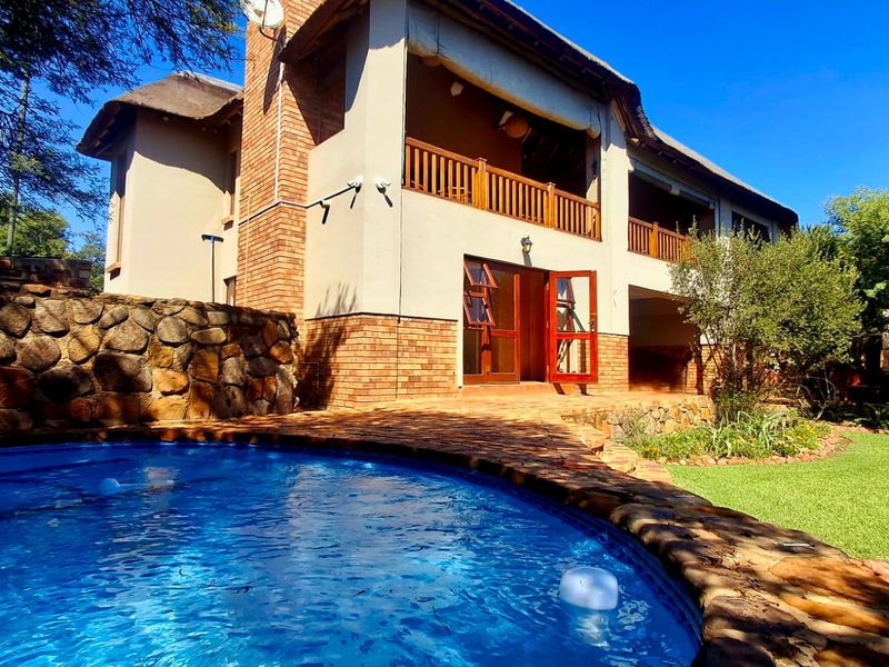 Rear view of a two-story house featuring a swimming pool, stone retaining wall, thatched roof, and a balcony with wooden railings.