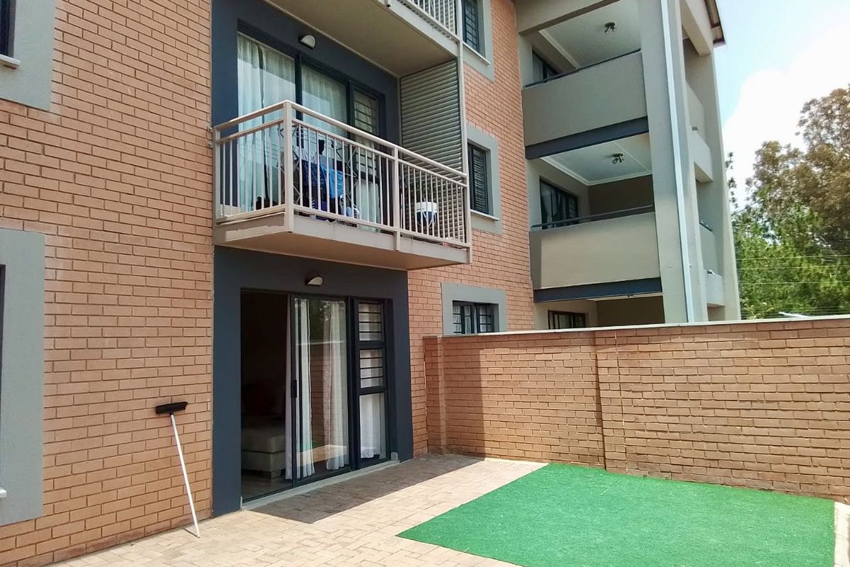 Rear view of the apartment building featuring a ground-floor paved patio with artificial grass, a brick boundary wall, and an upper-level balcony.