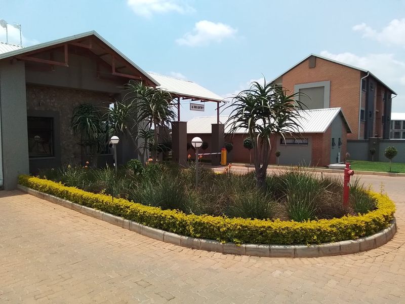 The entrance to the apartment complex features a paved driveway, landscaped garden beds, and a secure access boom gate.