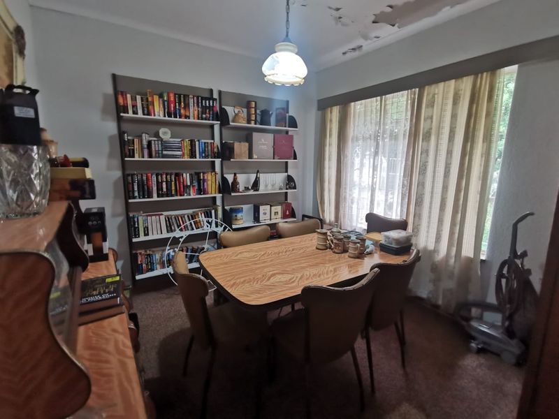 A dining room featuring built-in shelving, a central pendant light, and large windows, currently showing signs of wear on the ceiling and flooring.