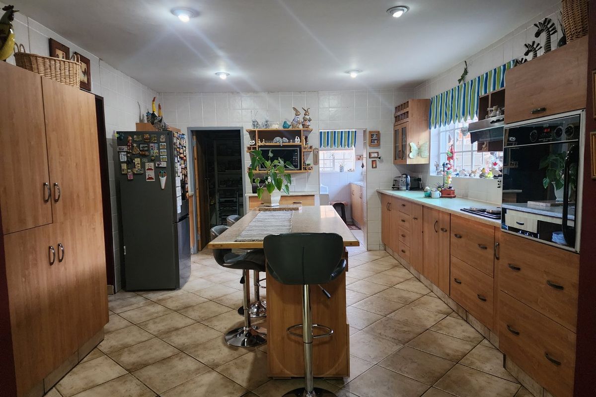 Spacious kitchen featuring wood-finish cabinetry, a central island with breakfast bar seating, built-in oven, and tiled flooring, leading to a separate scullery area.