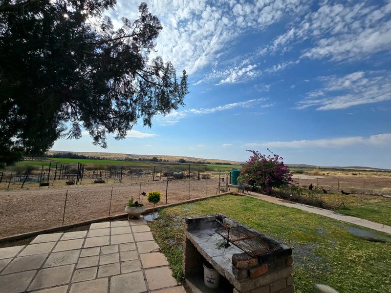Outdoor paved patio area featuring a built-in brick braai, overlooking a spacious rural garden and open landscape.