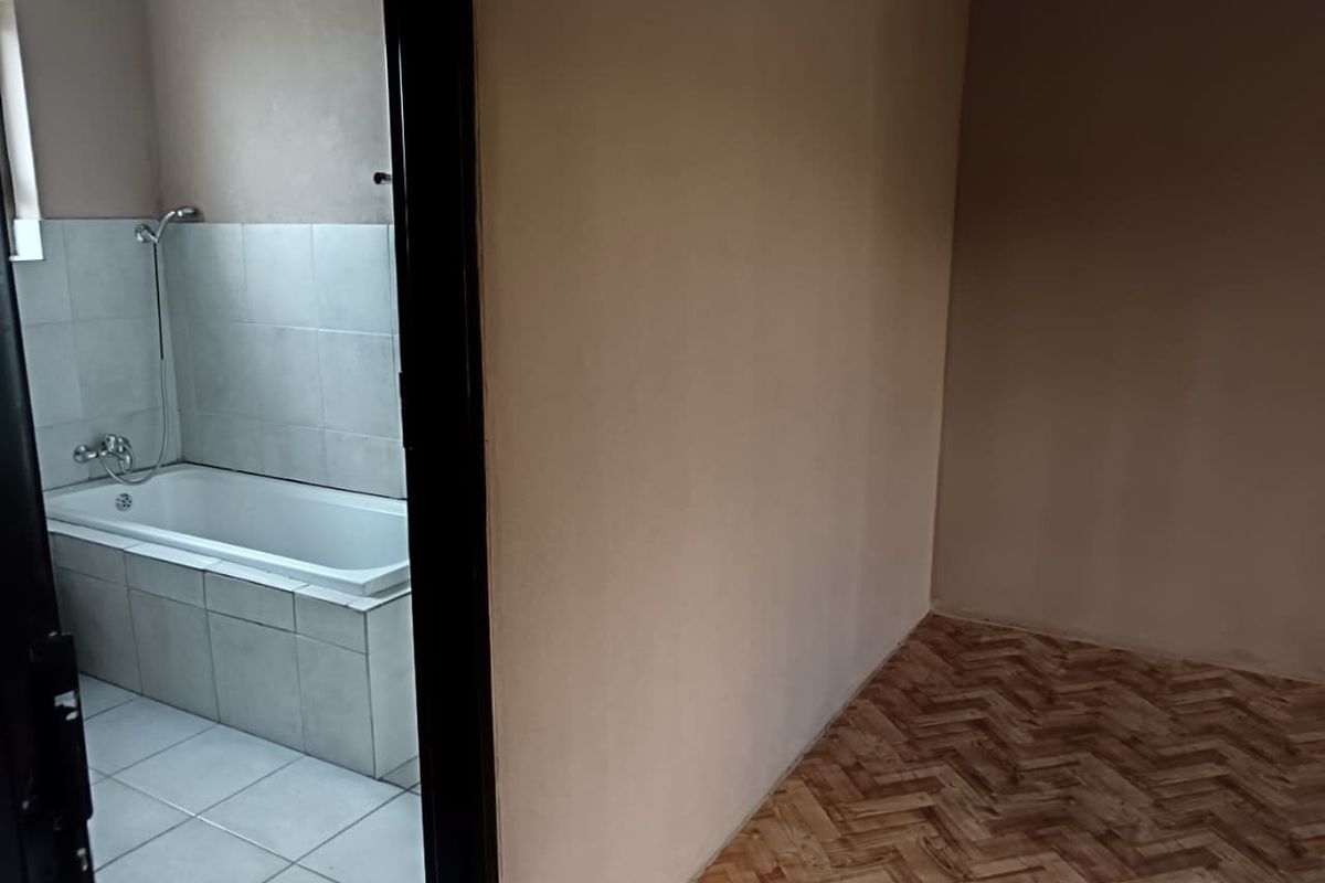 An empty room adjacent to a bathroom featuring a built-in bathtub with tiled surround and shower mixer.