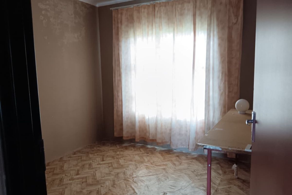 A vacant bedroom featuring parquet flooring and a window with curtain rails, showing visible signs of wall surface deterioration requiring maintenance.