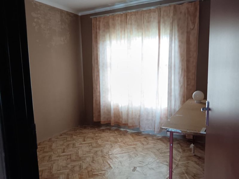 A vacant bedroom featuring parquet flooring and a window with curtain rails, showing visible signs of wall surface deterioration requiring maintenance.