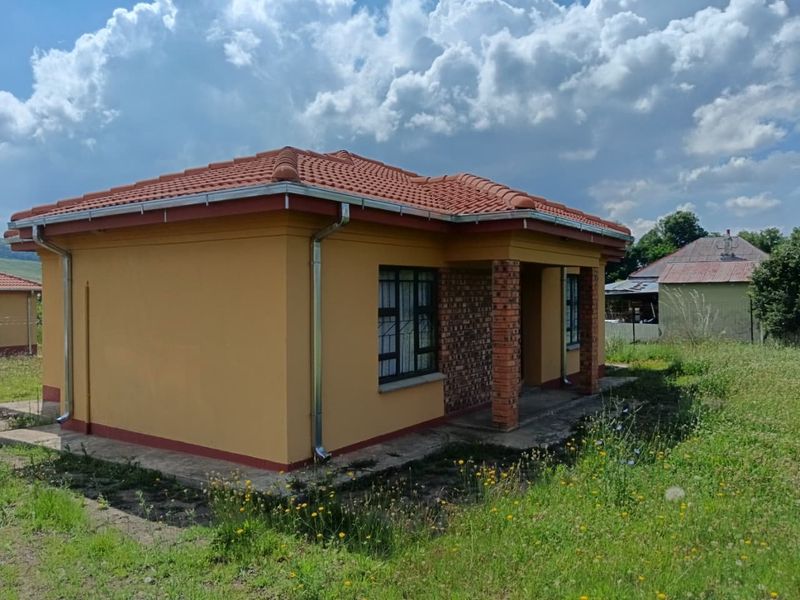 A single-story residential house featuring a tiled roof, brick pillar accents, and metal guttering, set on a plot with surrounding grass.