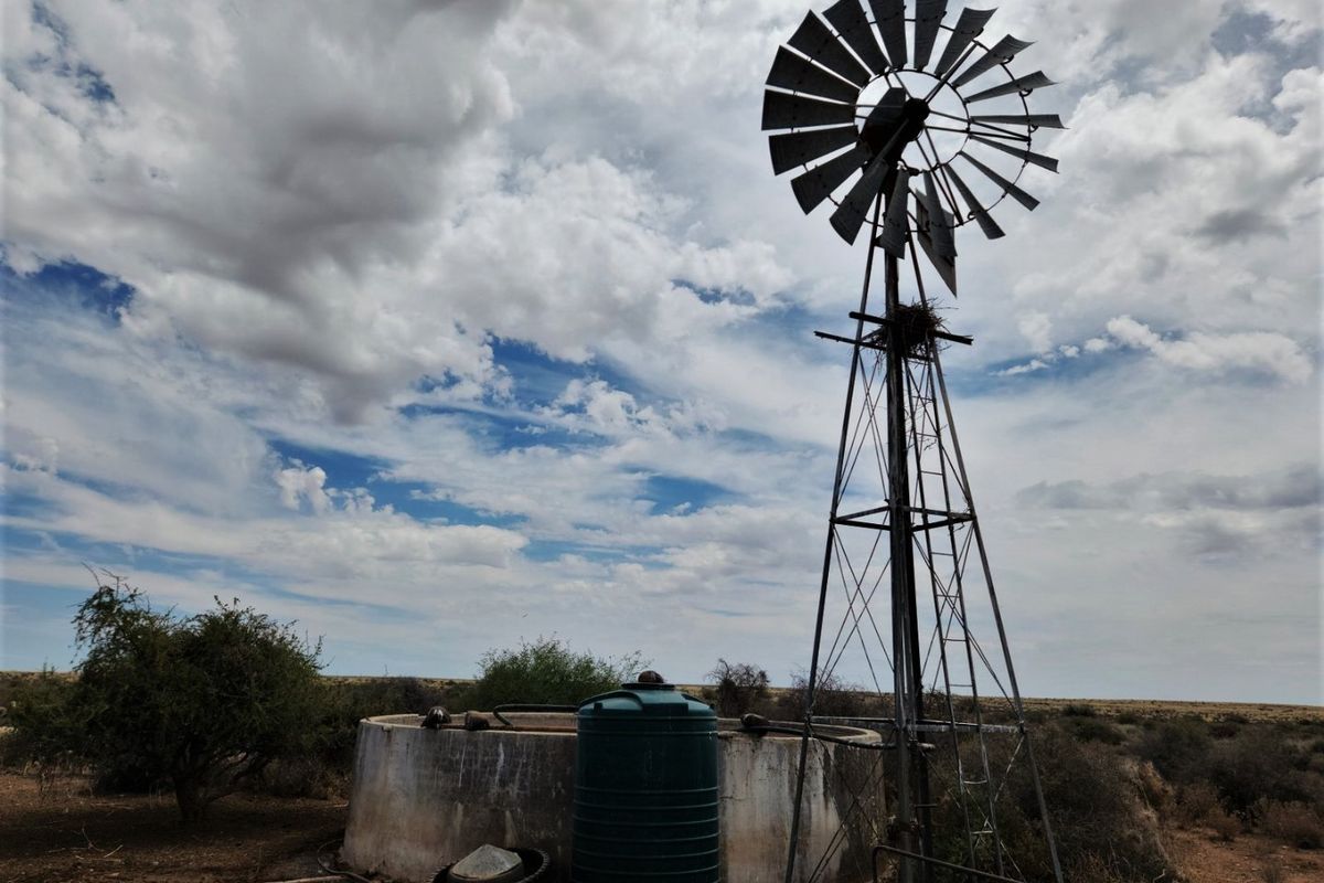 A rural farm utility area featuring a traditional metal windmill, a concrete water reservoir, and a plastic water storage tank.
