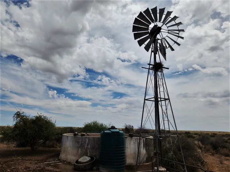 A rural farm utility area featuring a traditional metal windmill, a concrete water reservoir, and a plastic water storage tank.