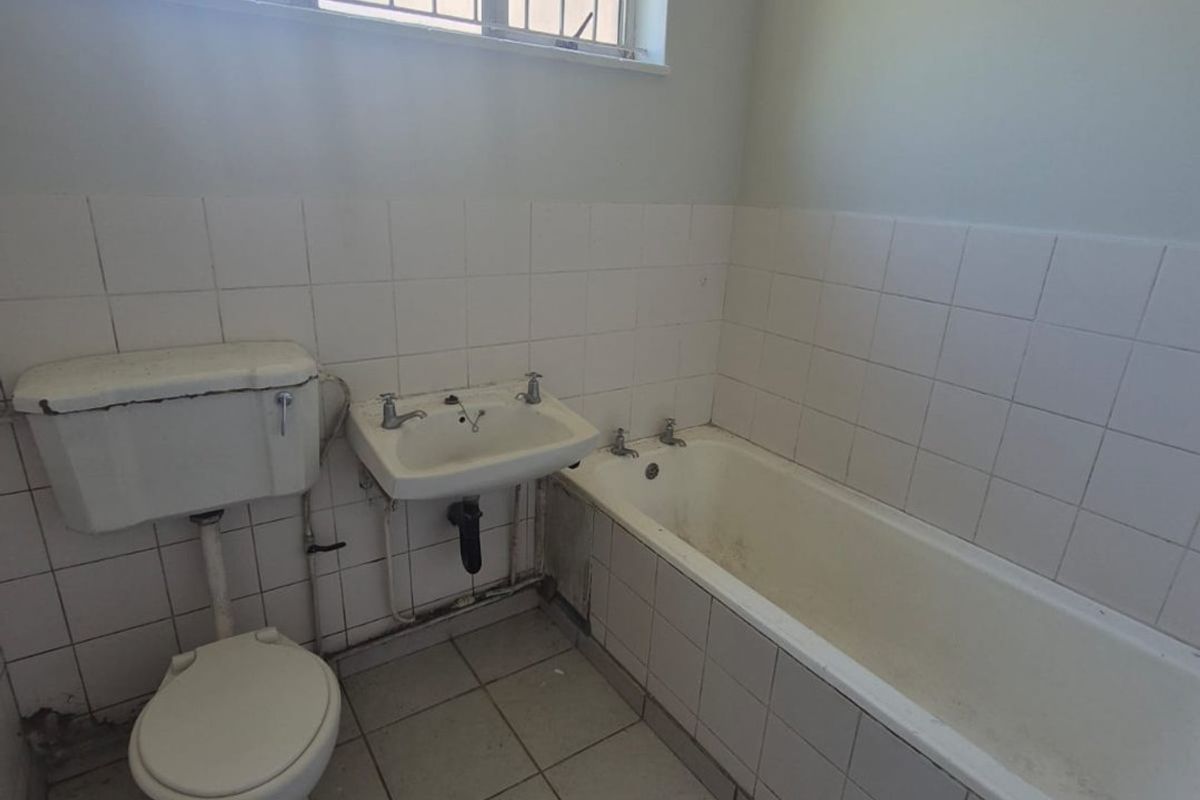 A dated bathroom featuring a standard bathtub, pedestal basin, and toilet, finished with white wall tiles and floor tiles, requiring cosmetic updates.