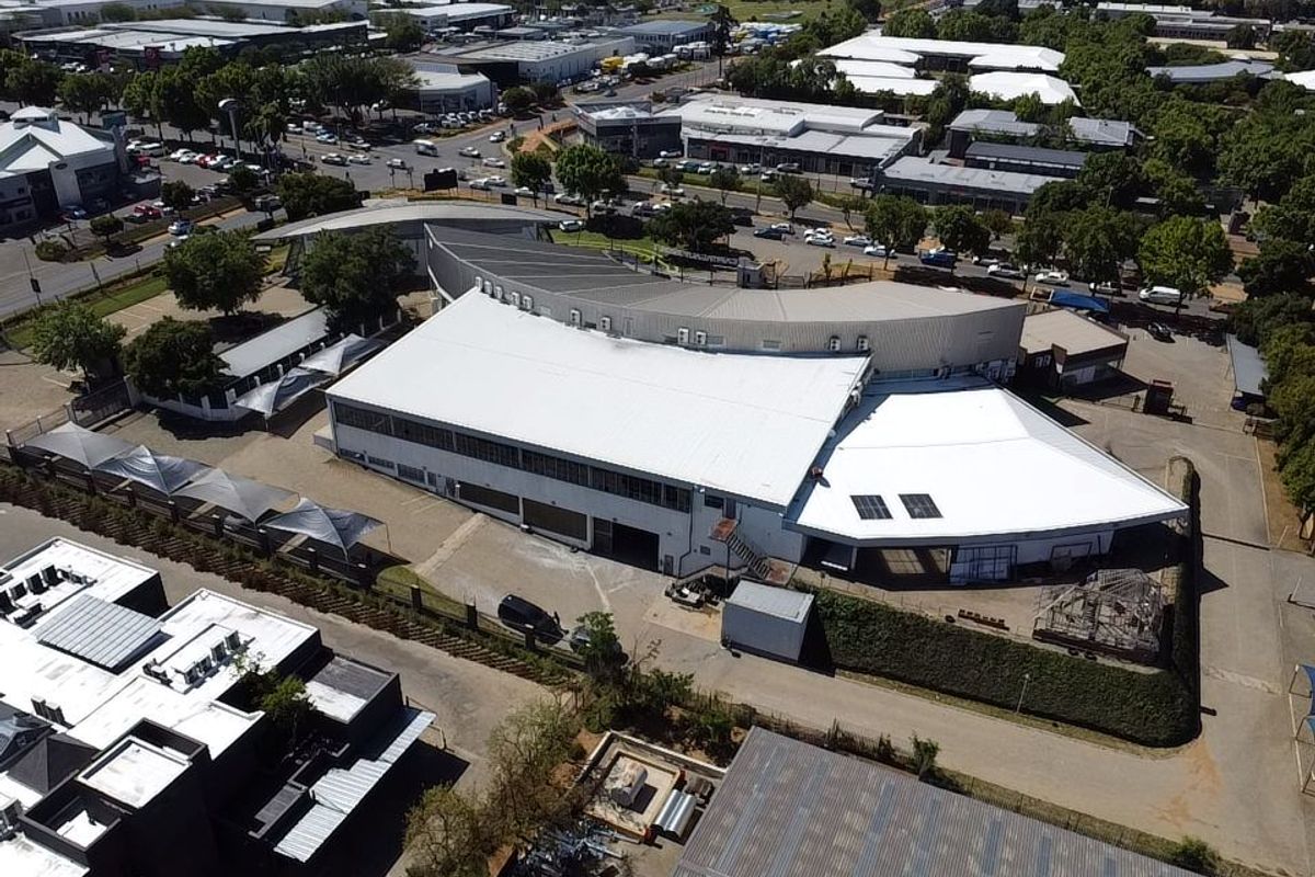 Aerial view of a commercial property featuring a large metal-roofed building, paved parking areas, and perimeter fencing within an established business park setting.
