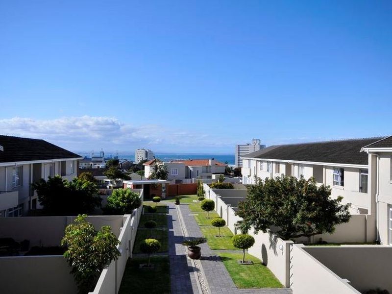 An elevated view of the apartment complex courtyard featuring landscaped garden paths, manicured trees, and surrounding residential units with a distant ocean view.