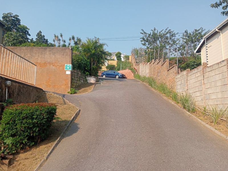 An upward sloping asphalt driveway leading to the property entrance, flanked by concrete boundary walls and security fencing.