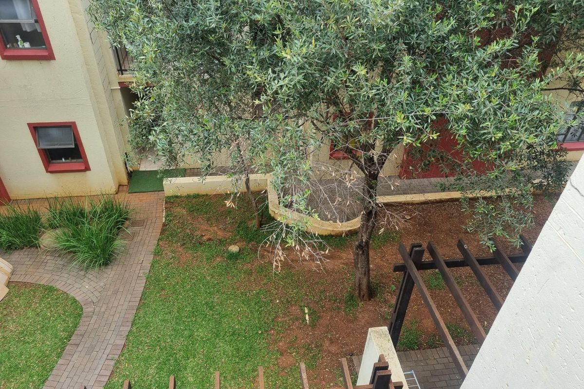An elevated view of a communal garden area featuring a paved walkway, a wooden pergola, and established trees within the apartment complex grounds.