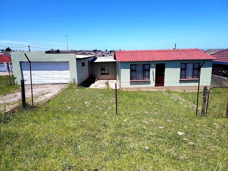 Front view of a single-story house featuring a garage, corrugated metal roof, and a large front yard with perimeter fencing.