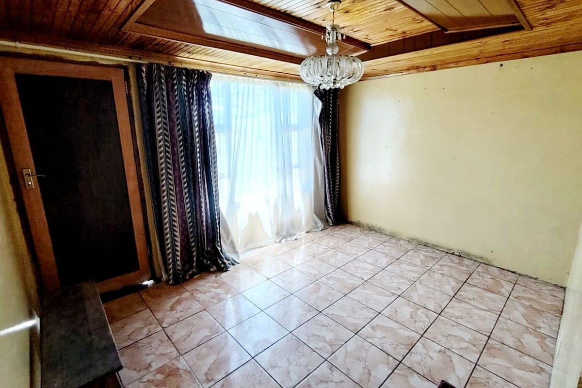 An empty living room featuring a timber-clad ceiling, ceramic floor tiles, and a crystal chandelier, requiring cosmetic updates to the walls.