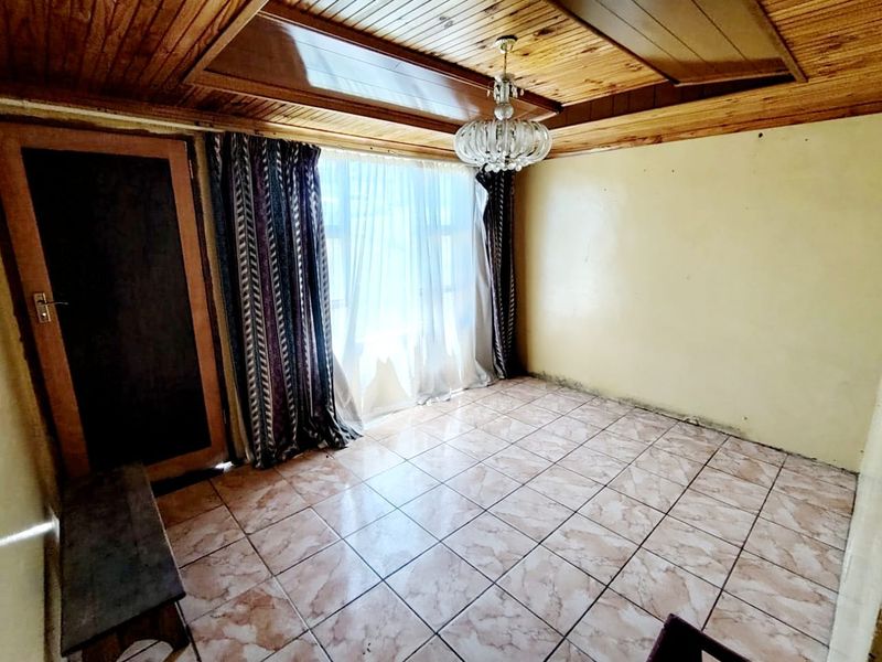 An empty living room featuring a timber-clad ceiling, ceramic floor tiles, and a crystal chandelier, requiring cosmetic updates to the walls.
