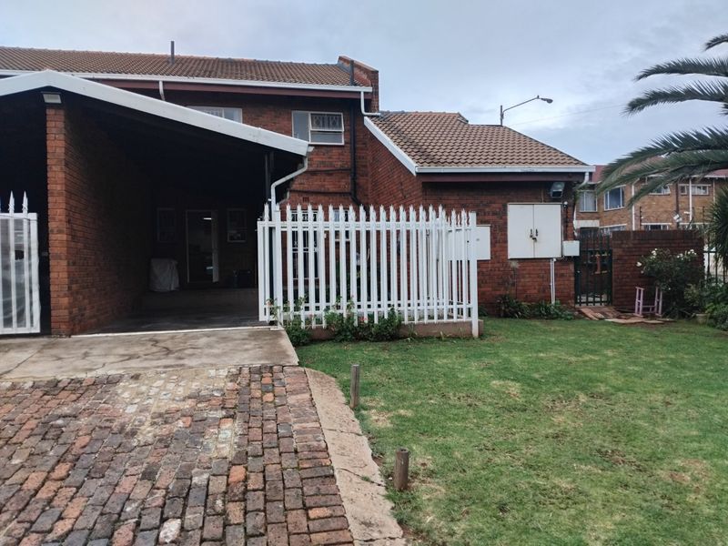 Front view of a brick apartment building featuring a covered carport, paved driveway, and a small private garden area with security fencing.