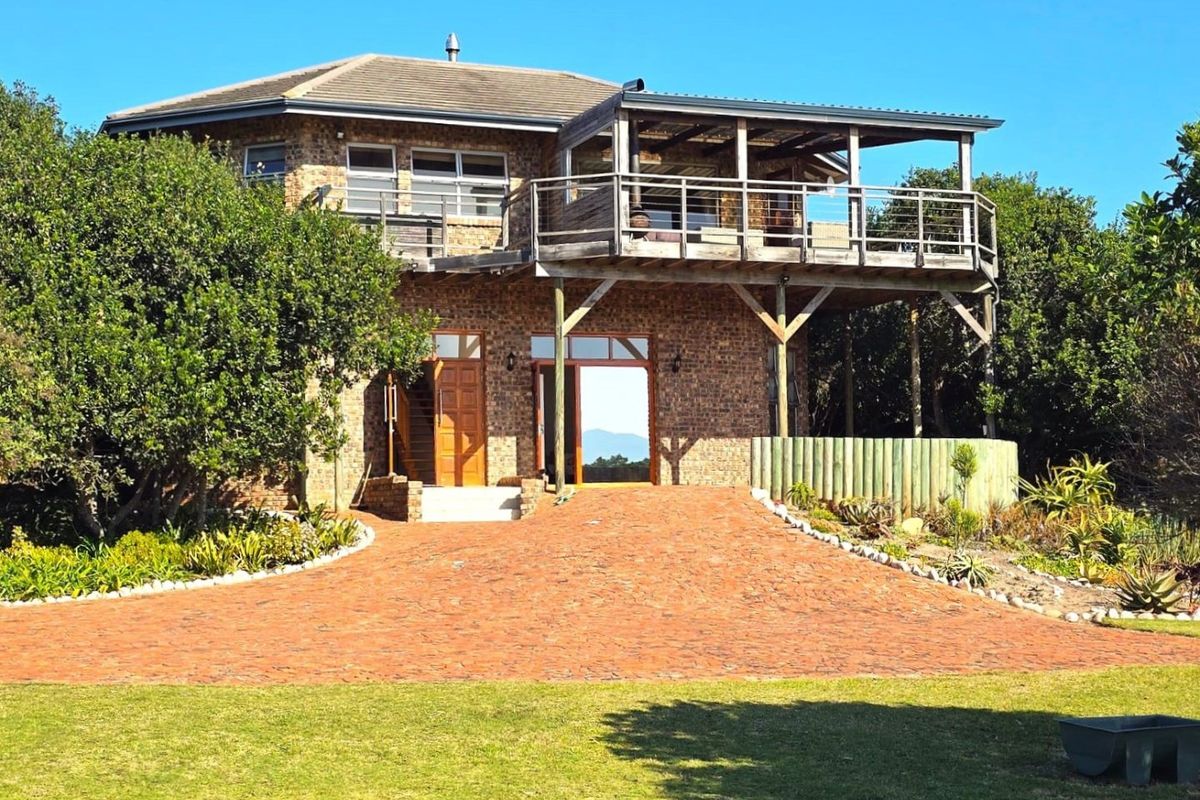 Rear view of a multi-level brick farmhouse featuring a large raised wooden deck, paved driveway, and established garden surroundings.