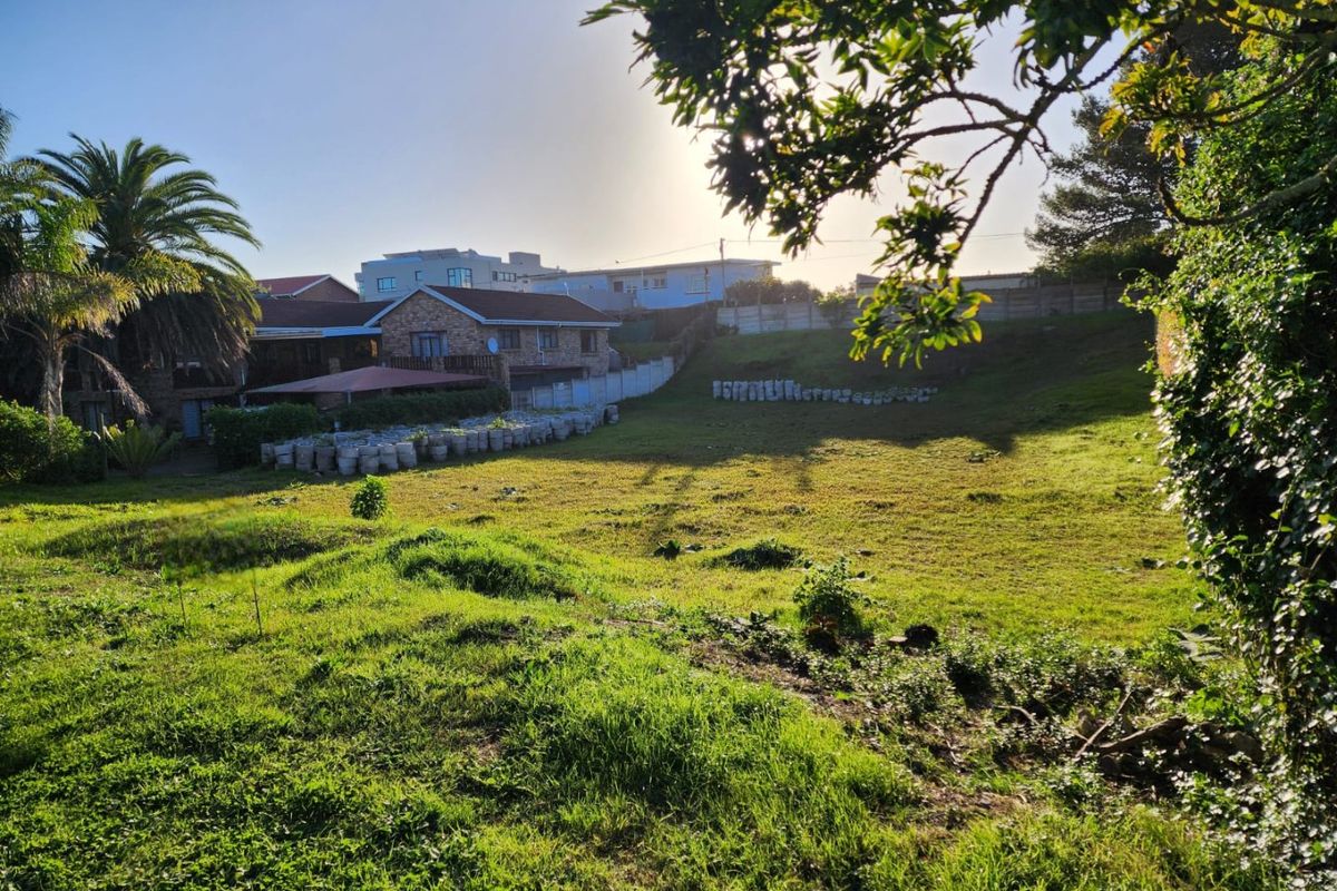 A view of a vacant residential plot featuring a grassy slope, perimeter fencing, and established trees, offering potential for future development.