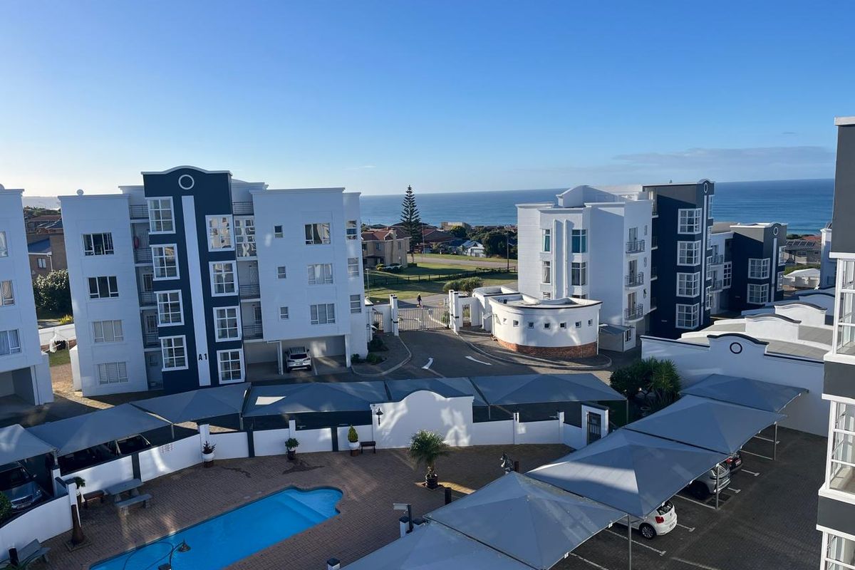 Elevated aerial view of the apartment complex featuring a swimming pool, covered parking bays, and secure gated access with ocean views in the distance.