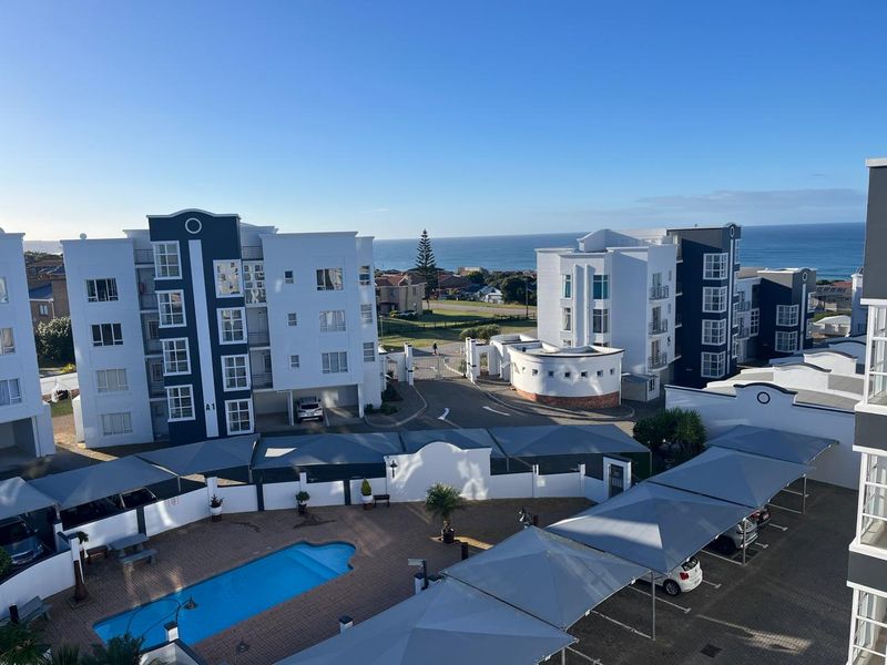 Elevated aerial view of the apartment complex featuring a swimming pool, covered parking bays, and secure gated access with ocean views in the distance.