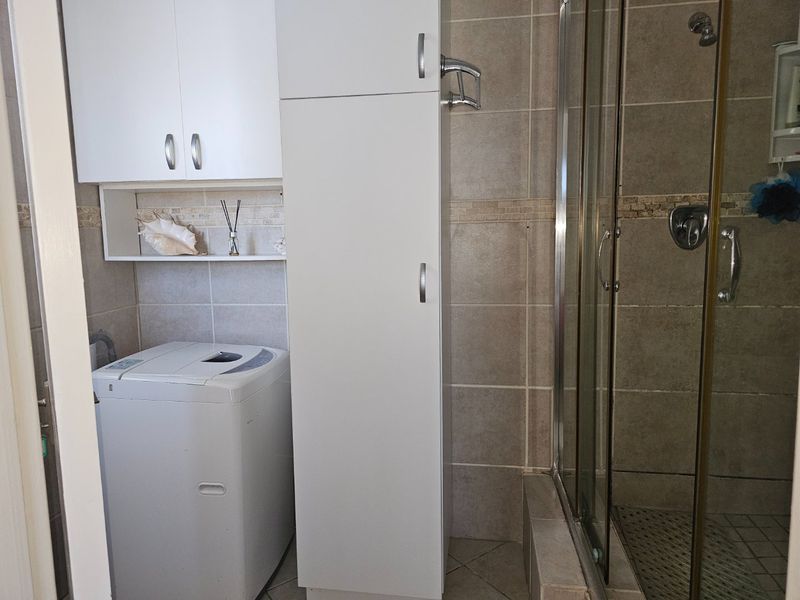 A functional laundry area integrated within a bathroom space, featuring built-in white cabinetry, tiled finishes, and a glass-enclosed shower.
