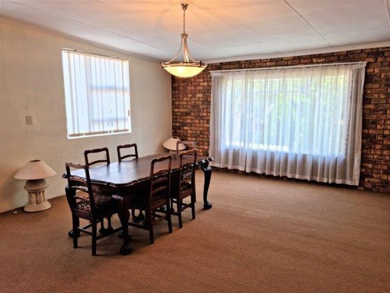 A dining room featuring an exposed brick feature wall, neutral carpeting, and a central pendant light fixture with large windows.