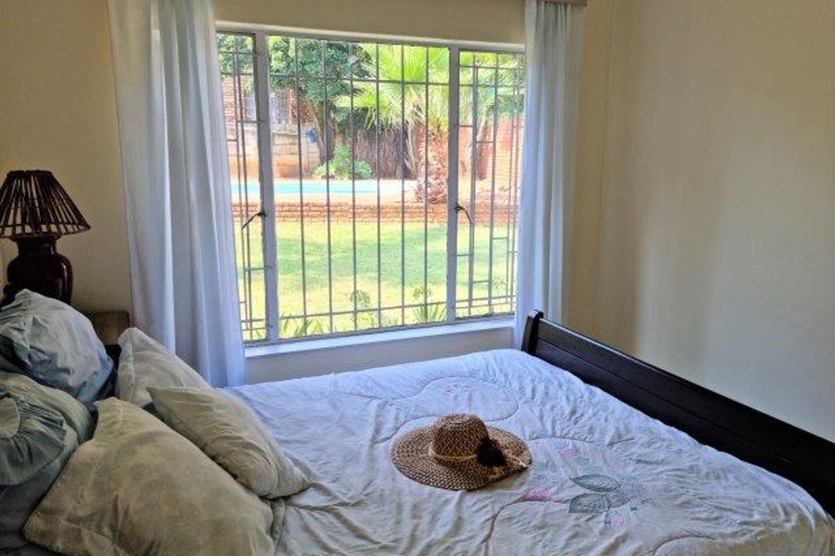 A bedroom featuring a large window with security bars, offering a view of the garden and pool area.